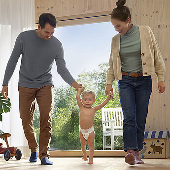 Eine Familie in einem Zimmer, mit einem Baby in einer Windel, das beide Eltern an den Händen halten, während sie zum Fenster gehen.